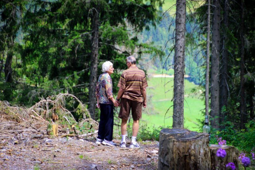 An older couple, holding hands and looking at each other in a forest.