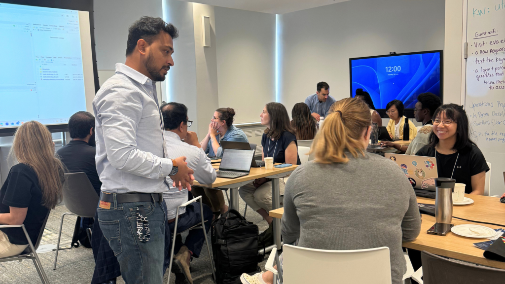 Sangram chatting with two workshop attendees at their table. Jeff is seen in the back supporting another table of attendees.